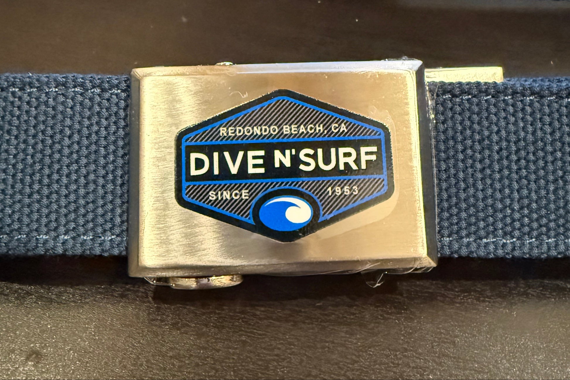 A navy blue woven web belt with a polished silver-tone buckle featuring a blue and white 'Dive N'Surf' logo, displayed on a dark tabletop.