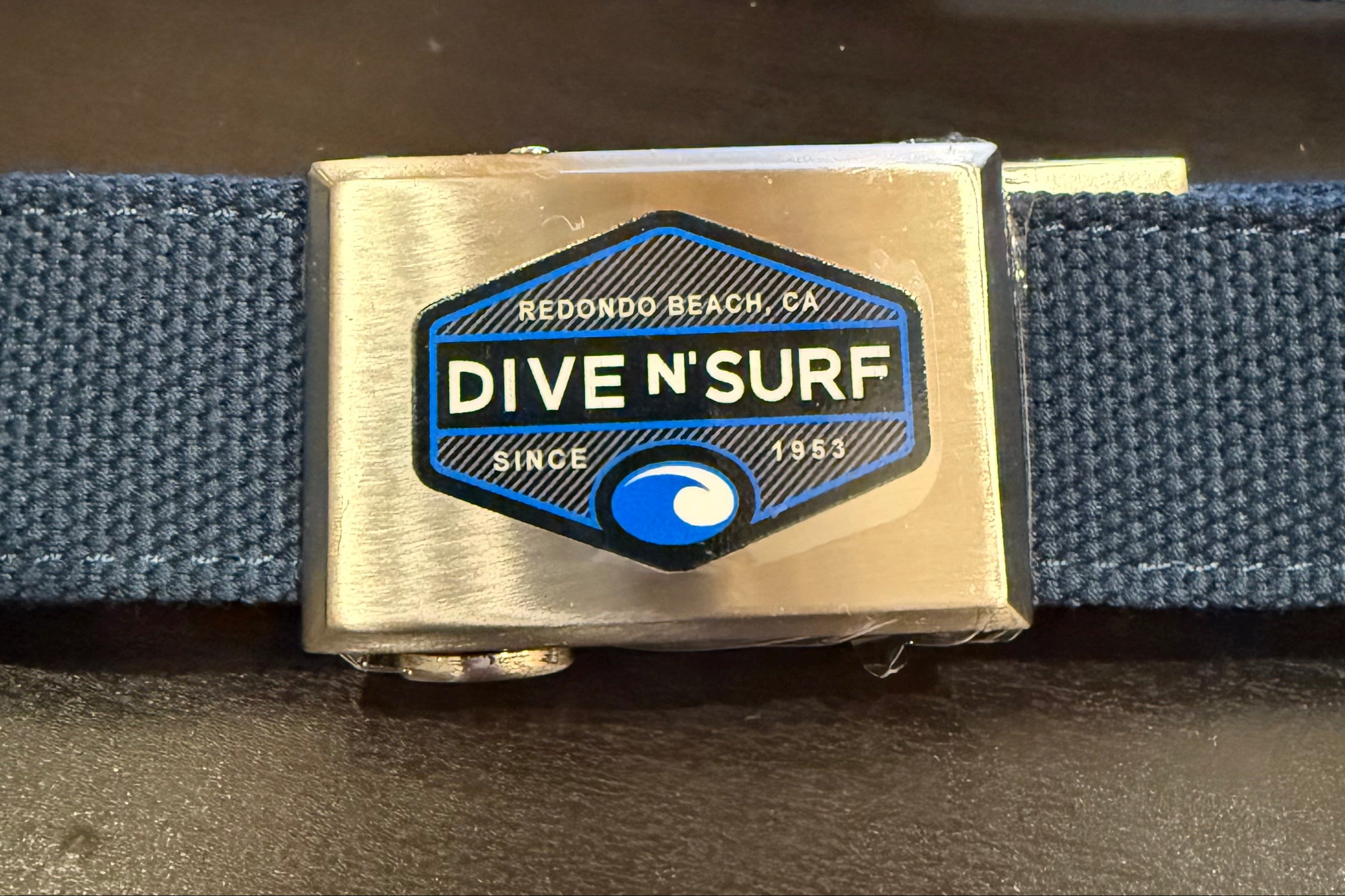 A navy blue woven web belt with a polished silver-tone buckle featuring a blue and white 'Dive N'Surf' logo, displayed on a dark tabletop.